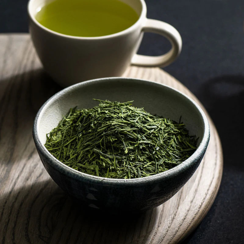 Thé vert Gyokuro bio en feuilles entières dans un bol en céramique, avec tasse de thé infusé à l’arrière-plan.
